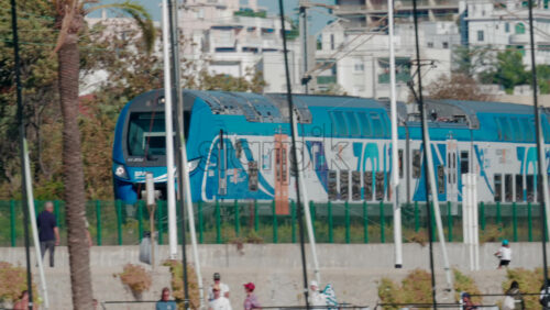 Video - A modern blue regional train passes along the seaside railway, with palm trees and people in the foreground