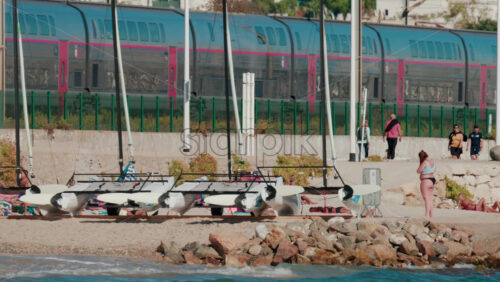 Video - Cannes, France - October 6, 2025: A modern train passes along the coastline near the beach, while people walk and relax nearby