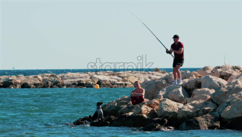 Video - Cannes, France - October 6, 2025: A man fishing from the rocky shore while a woman and a dog sit nearby, enjoying the calm sea in Cannes, France