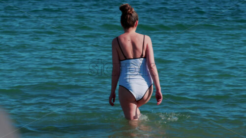 Video - A young woman walks slowly into the calm turquoise water on a sunny summer day in Cannes, France