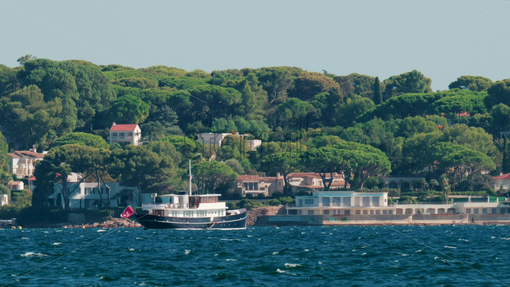 Video - A stylish black yacht sails along the Cannes coastline surrounded by pine trees and villas under clear blue skies on the French Riviera