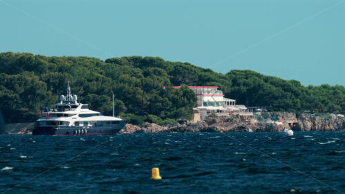 Video - A luxurious white yacht anchored near a rocky Mediterranean coastline surrounded by lush green pine trees and elegant seaside villas