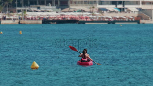 Video - Two women paddle a red kayak across calm turquoise waters near a sunny beach resort
