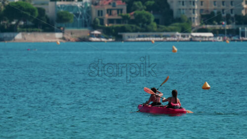 Video - Two women paddle a red kayak across calm turquoise waters near a sunny beach resort