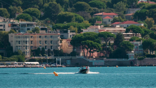 Video - A sleek motorboat speeds across the clear blue water with coastal buildings and lush trees in the background