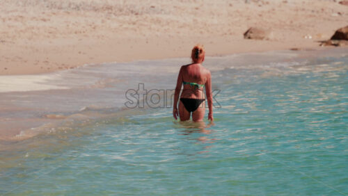 Video - A woman in a bikini walks slowly into the clear turquoise water along a sandy beach under the sun