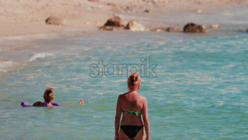 Video - A woman in a bikini walks slowly into the clear turquoise water along a sandy beach under the sun