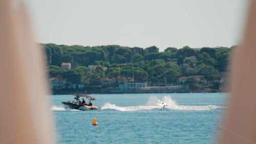 Video - A wakeboarder glides over the waves behind a speedboat on a sunny coastal day, with scenic villas and pine covered hills in the background