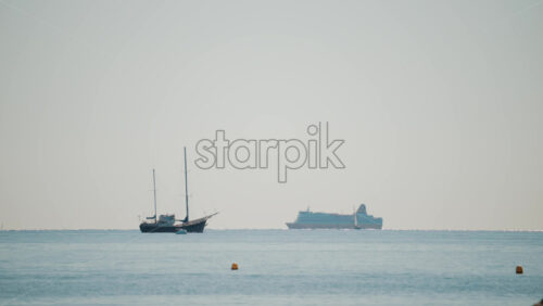 Video - Wide shot of a calm sea with a sailboat and a large cruise ship sailing in the distance under a pale blue sky
