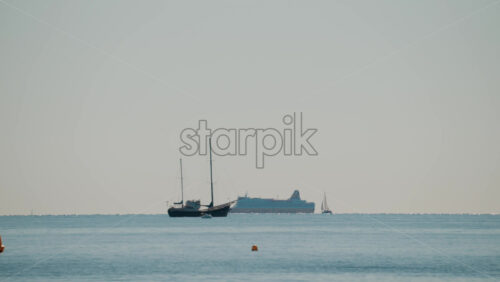 Video - Wide shot of a calm sea with a sailboat and a large cruise ship sailing in the distance under a pale blue sky