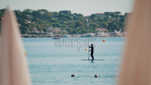 Video - A man stand-up paddleboarding on tranquil blue water near a coastal town, framed by blurred umbrellas in the foreground