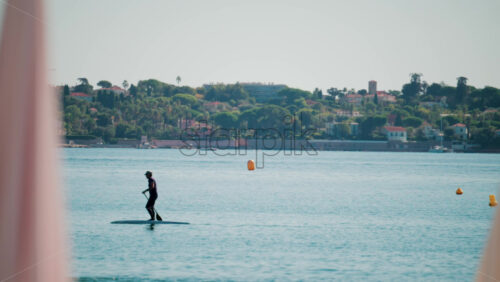 Video - A man stand-up paddleboarding on tranquil blue water near a coastal town, framed by blurred umbrellas in the foreground