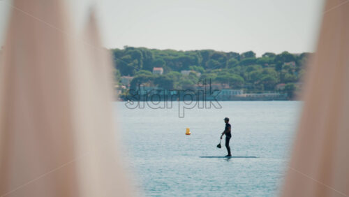 Video - A man stand-up paddleboarding on tranquil blue water near a coastal town, framed by blurred umbrellas in the foreground
