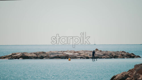 Video - A man on a paddleboard approaches a rocky breakwater on a bright sunny day