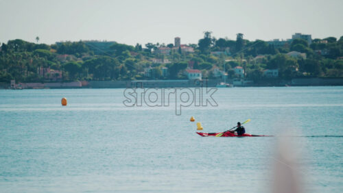 Video - A man paddles a red kayak across the calm turquoise sea near a coastal town