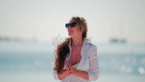 Video - Smiling woman wearing sunglasses and a white shirt stands on the beach under the sun, with the sea glittering behind her
