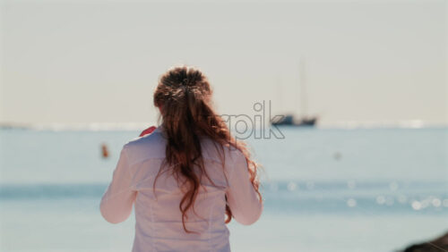 Video - Smiling woman wearing sunglasses and a white shirt stands on the beach under the sun, with the sea glittering behind her