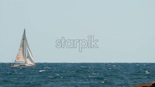 Video - Cannes, France - October 7, 2025: A white sailboat navigates through blue waves on a clear sunny day, with the horizon stretching endlessly behind it
