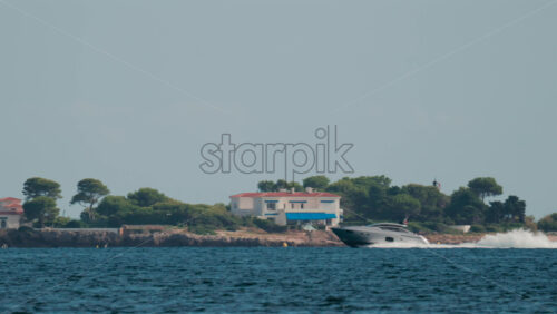 Video - A fast moving yacht glides past a luxury coastal villa surrounded by trees and clear blue water