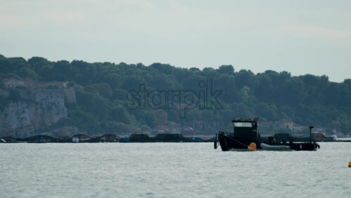 Video - A black industrial boat floating beside offshore fish farming nets with forested hills in the background