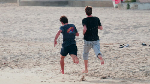 Video - Two young men run barefoot across the sand on a sunny day, enjoying outdoor exercise and friendship