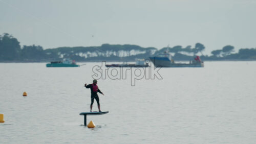 Video - A person rides an electric foilboard above calm blue water, wearing a helmet and life vest