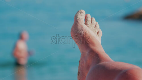 Video - Close up of sandy feet resting with the turquoise sea in the background