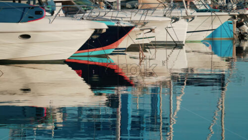 Video - Close up of several yachts moored in a marina, with vivid reflections of their hulls on calm water