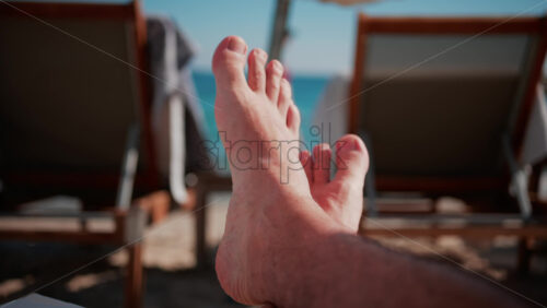 Video - Close up of bare feet resting on a beach lounger with soft sunlight and blurred deck chairs in the background
