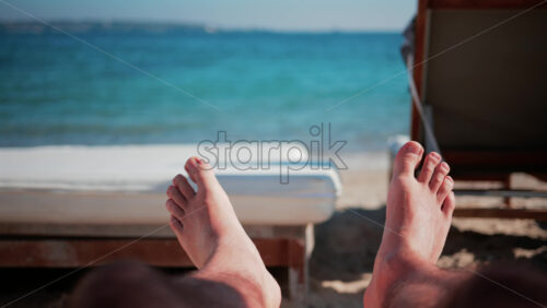 Video - Close up of bare feet resting on a beach lounger with soft sunlight and blurred deck chairs in the background