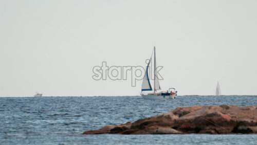 Video - A sailboat glides in the distance behind rocky coastal formations