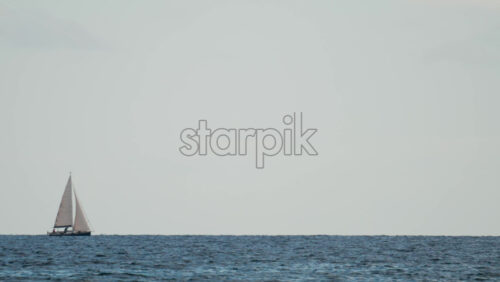 Video - A white sailboat navigates through blue waves on a clear sunny day, with the horizon stretching endlessly behind it