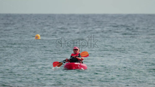 Video - Cannes, France - October 8, 2025: A woman and a man paddling a red kayak on calm blue water wearing a life vest and hat
