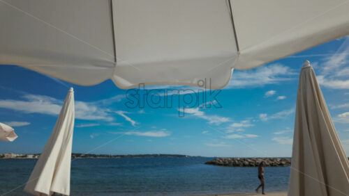Video - Wide shot of a man walking along the beach under large white umbrellas with a clear blue sky in the background