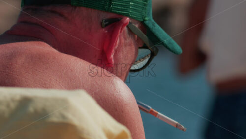 Video - Cannes, France - October 10, 2025: Close up of an elderly man wearing a green cap and sunglasses, smoking a cigarette while sitting by the sea