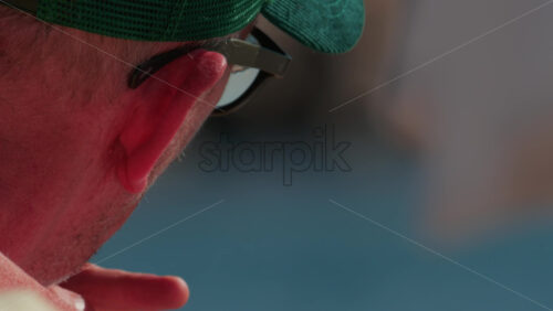Video - Cannes, France - October 10, 2025: Close up of an elderly man wearing a green cap and sunglasses, sitting by the sea