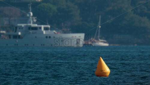Video - A bright yellow buoy floating on calm blue water with a yacht blurred in the background