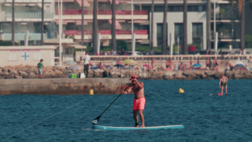 Video - Cannes, France - October 10, 2025: A man in bright swim shorts paddles near the shore with beachgoers and palm lined buildings in the background