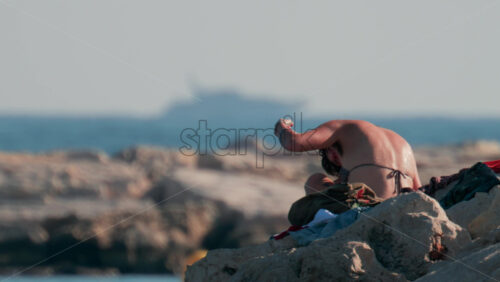 Video - Cannes, France - October 10, 2025: A young woman with wet hair relaxes on seaside rocks under the warm sun, with the sea and a yacht in the distance