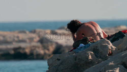 Video - Cannes, France - October 10, 2025: A young woman with wet hair relaxes on seaside rocks under the warm sun, with the sea and a yacht in the distance