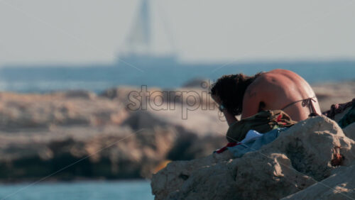 Video - Cannes, France - October 10, 2025: A young woman with wet hair relaxes on seaside rocks under the warm sun, with the sea and a yacht in the distance