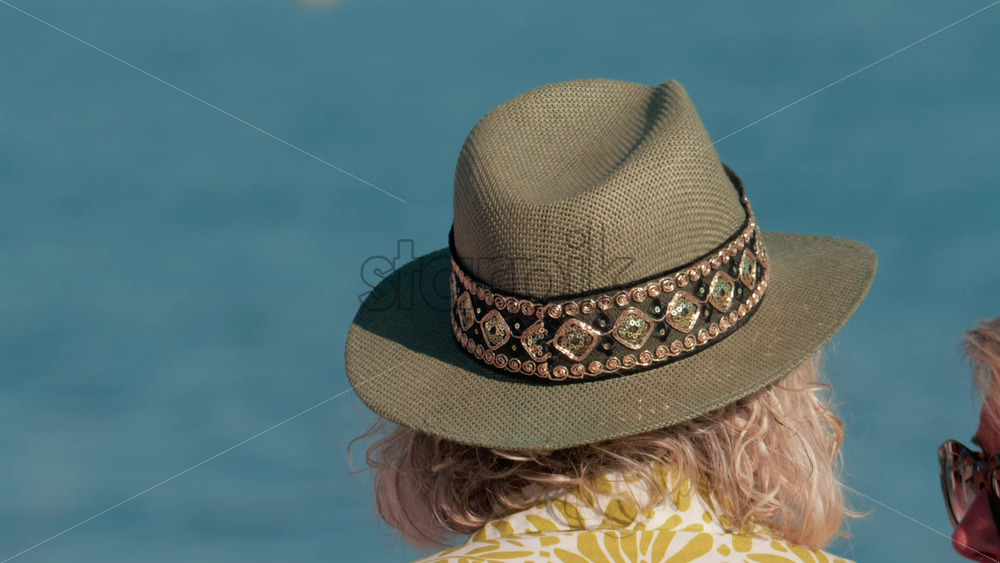 Video - Close up of a decorated sun hat worn by a person facing the calm blue water