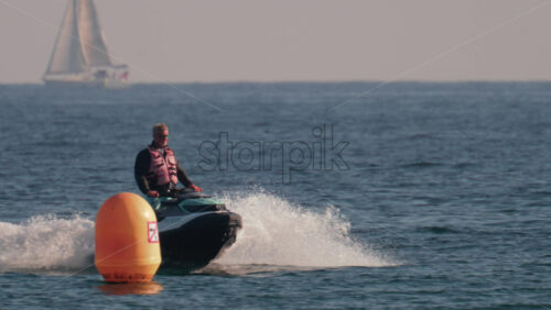 Video - Cannes, France - October 10, 2025: A man in a life jacket rides a jet ski past an orange buoy with sailboats in the background
