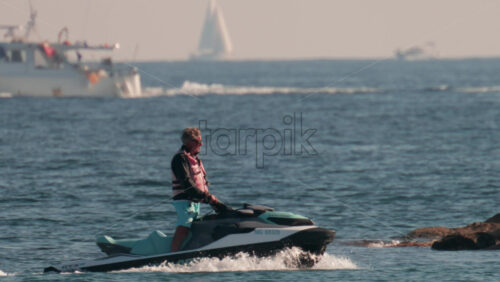 Video - Cannes, France - October 10, 2025: A man in a life jacket rides a jet ski past an orange buoy with sailboats in the background