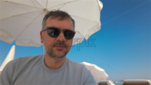 Video - Portrait of a man wearing sunglasses under beach umbrellas with palm trees and a bright blue sky in the background