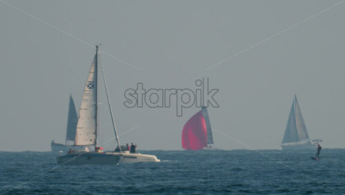 Video - Cannes, France - October 10, 2025: Several sailboats and a catamaran glide over the open sea under a hazy sky, with one striking red sail standing out