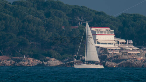Video - Cannes, France - October 10, 2025: A white sailboat glides near the rocky coastline of the French Riviera, with lush pine trees and elegant seaside villas in the background