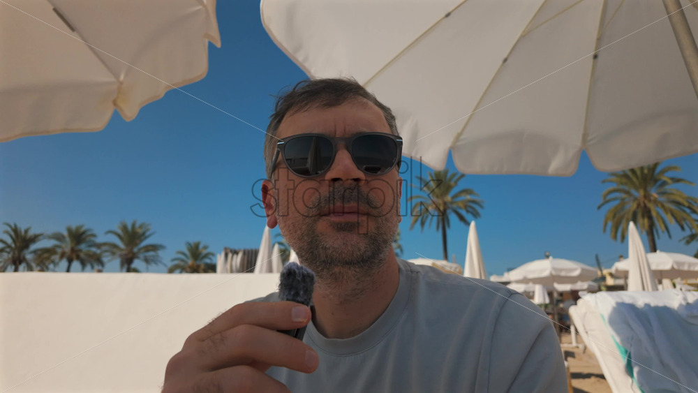 Video - Man wearing sunglasses speaks into a small microphone under large white beach umbrellas with palm trees in the background