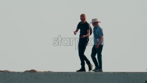 Video - Cannes, France - October 11, 2025: Two men walk side by side along a seaside pier under clear skies, engaged in relaxed conversation