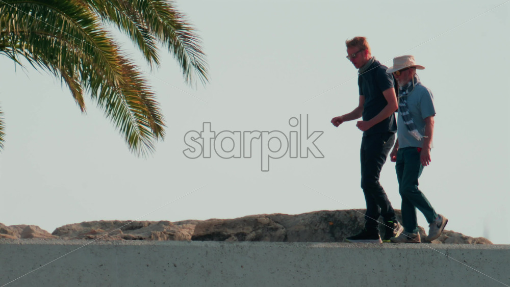 Video - Cannes, France - October 11, 2025: Two men walk side by side along a seaside pier under clear skies, engaged in relaxed conversation